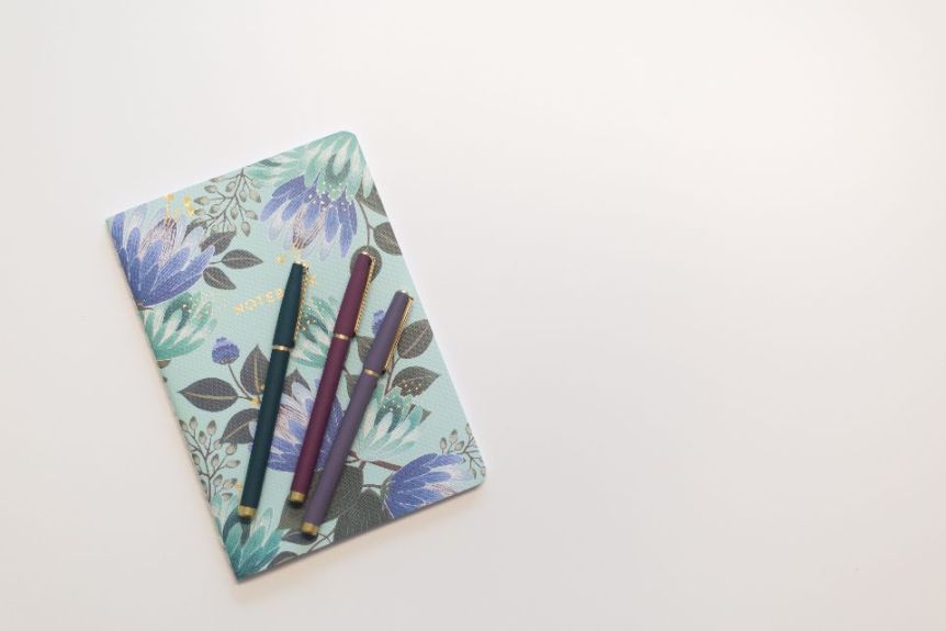 A floral notebook with three pens on a pale desk
