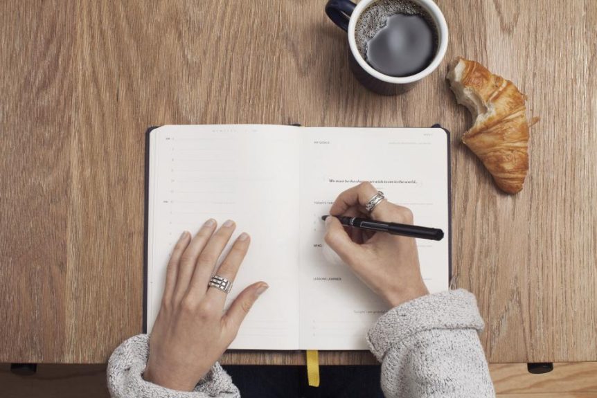 Hands writing in an open journal on a wooden table next to coffee and a croissant