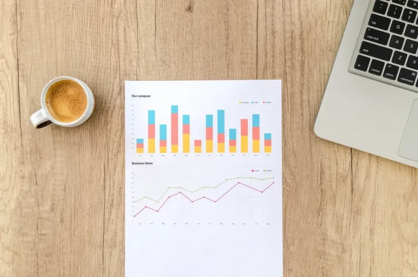 Printed bar and line charts on a wooden desk beside a laptop and a cup of coffee