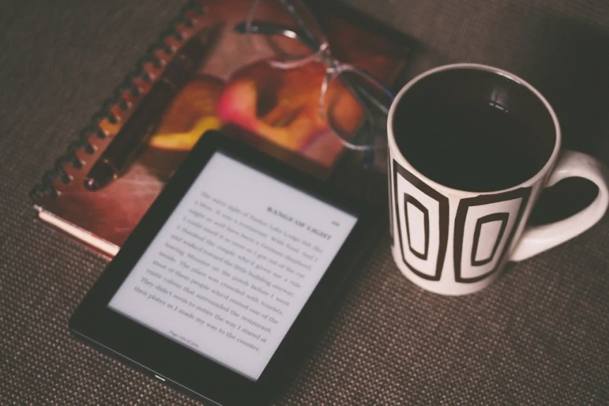 E-reader showing a page of text beside a coffee mug and notebook on a table