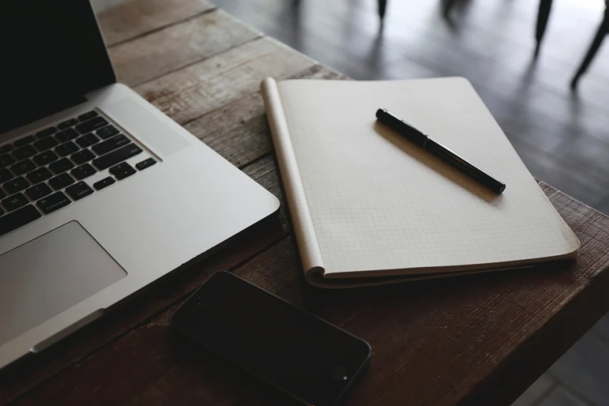 Open laptop next to a blank notebook and pen on a wooden desk