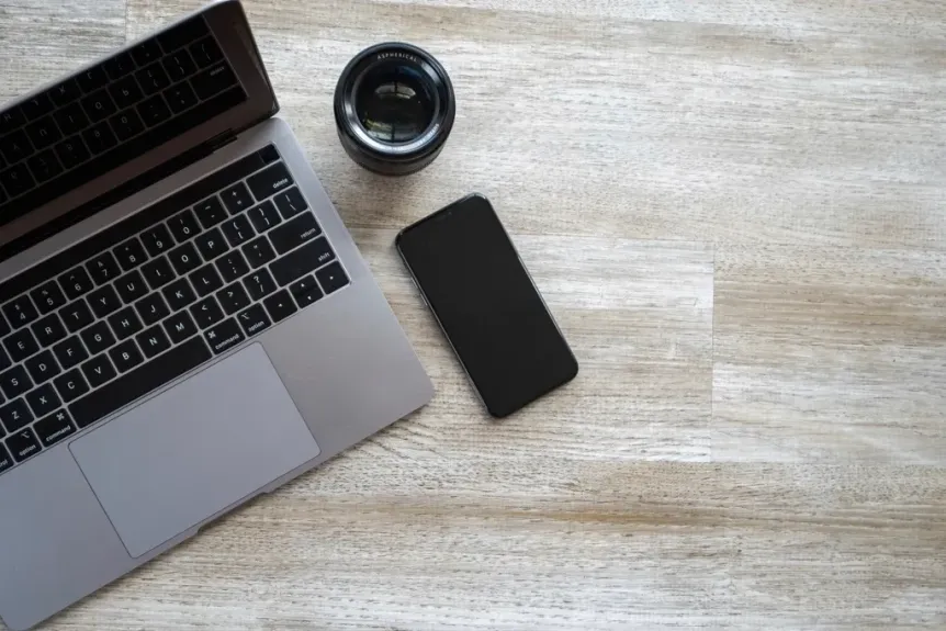 Overhead view of a laptop, smartphone and camera lens on a light wooden desk