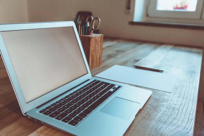 Open laptop on a wooden desk beside a sheet of paper and a pen