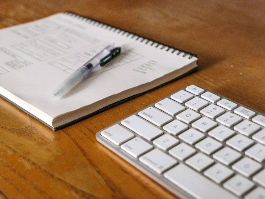 Spiral notebook with printed notes and a pen beside a white keyboard on a wooden desk