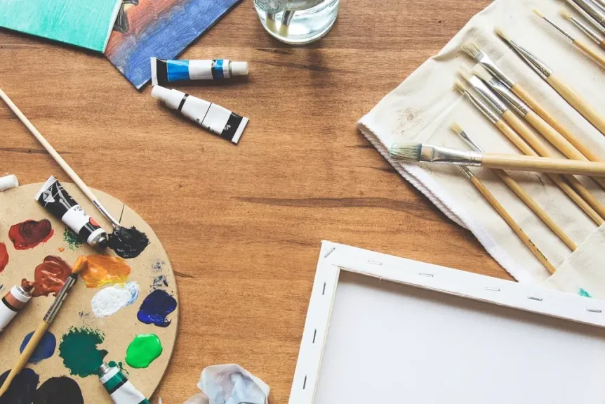 Paint brushes, paint tubes, a palette and a blank canvas on a wooden table