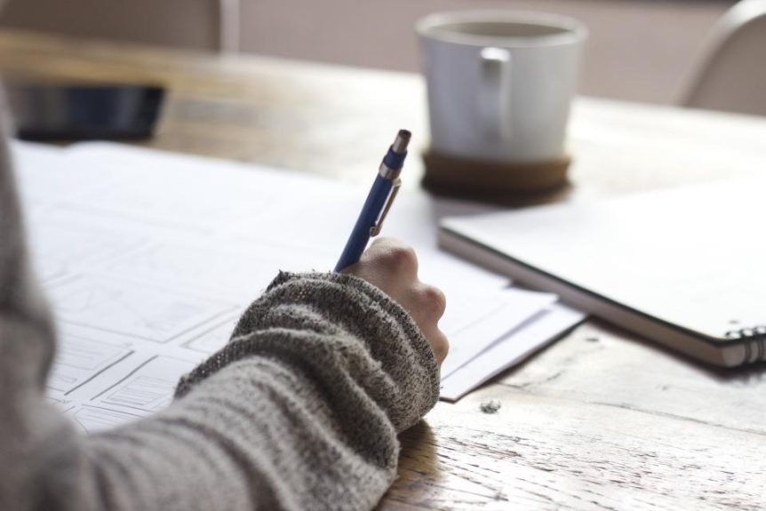 Hand writing in a notebook beside a coffee mug